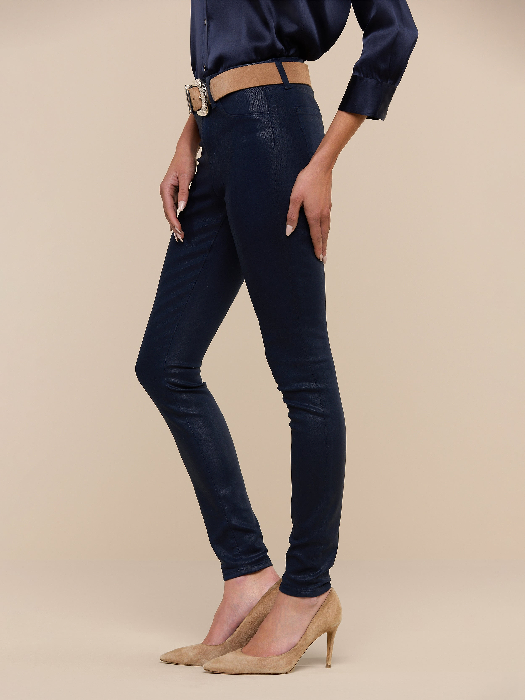 A person wearing the Marguerite Coated Skinny Jean in navy with a tapered leg, paired with a matching navy blouse, a tan belt, and tan high heels stands against a plain white background.