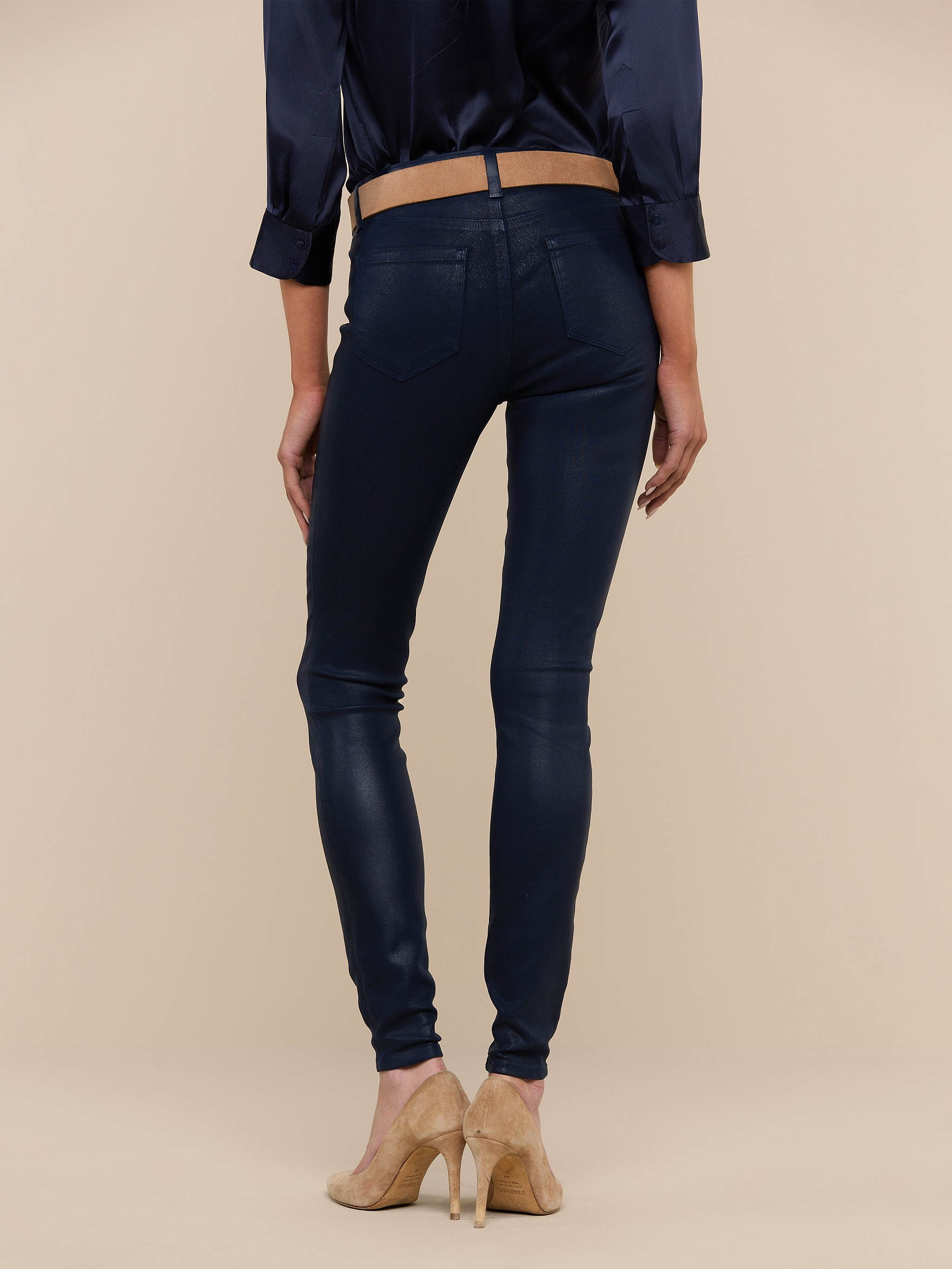 A person stands facing away, wearing a navy blouse and the Marguerite Coated Skinny Jean in navy with a tapered leg, paired with beige high-heeled shoes against a plain white background.