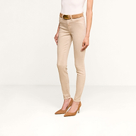 베이지색 ‘마르그리트 스키니 진’을 입고, 흰색 민소매 상의와 탄색 벨트, 탄색 하이힐을 신은 한 사람이 단색 베이지색 배경 앞에 서 있다.