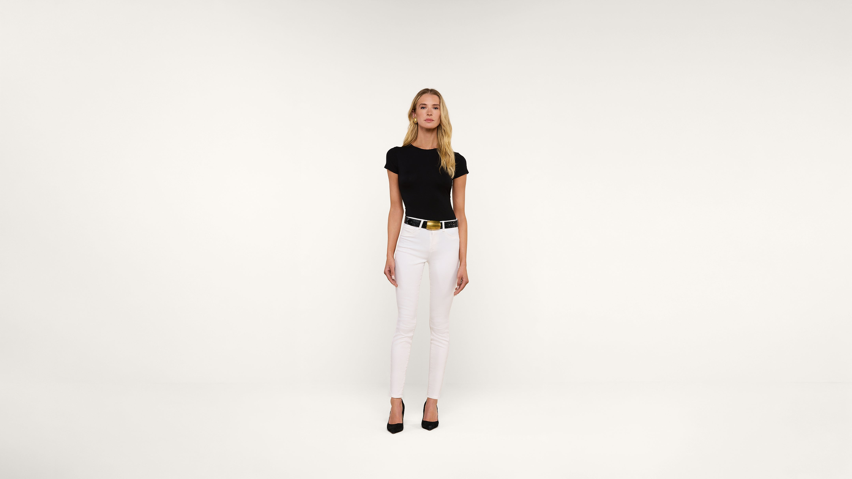 A woman stands facing forward against a plain white background, wearing a black t-shirt, white pants, a black belt with a gold buckle, and black heels.