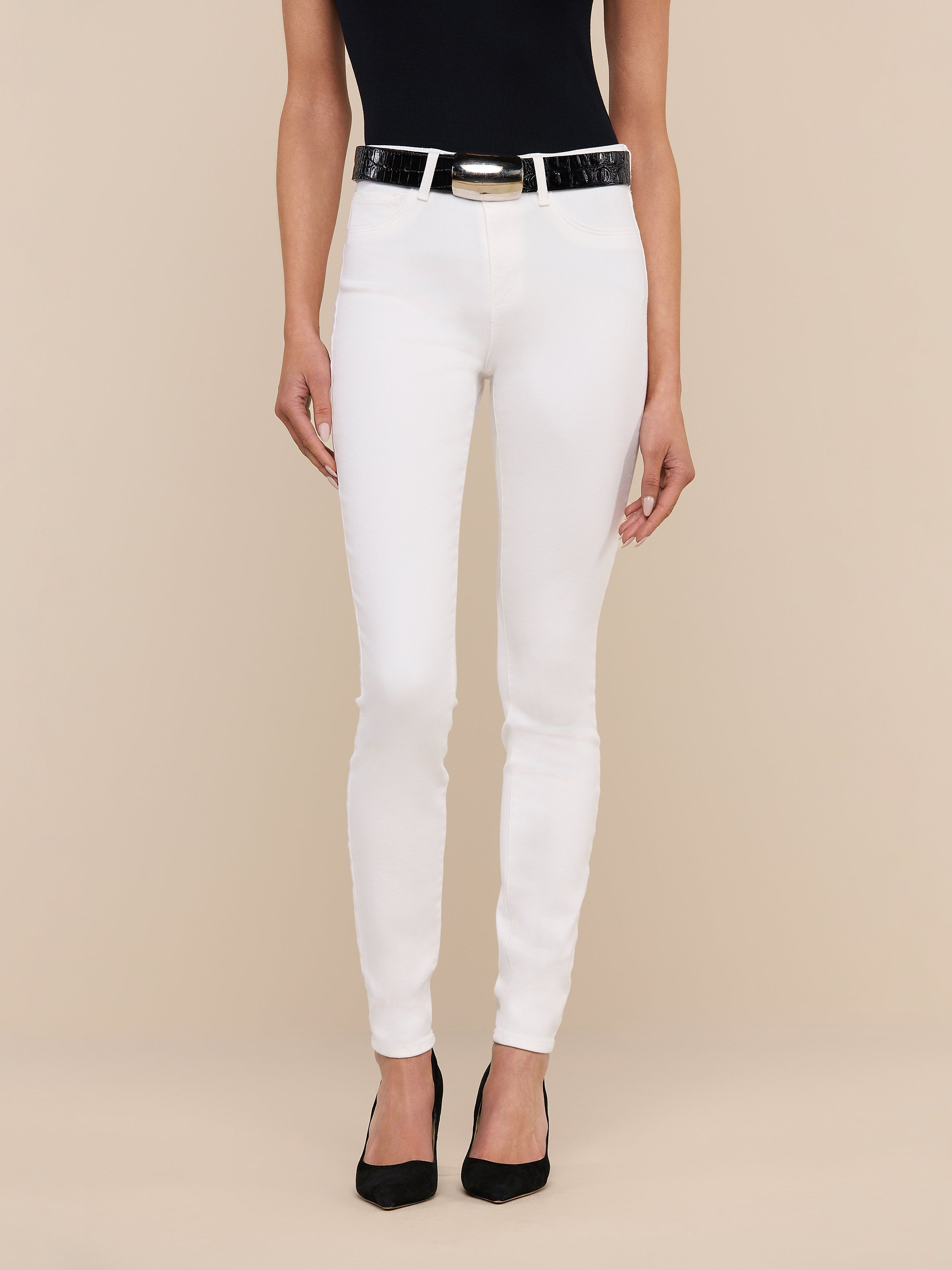 A woman stands against a plain background, wearing a black sleeveless top, the Marguerite Skinny Jean in white with a black belt, and black pointed-toe shoes. Only her body from the shoulders down is visible.