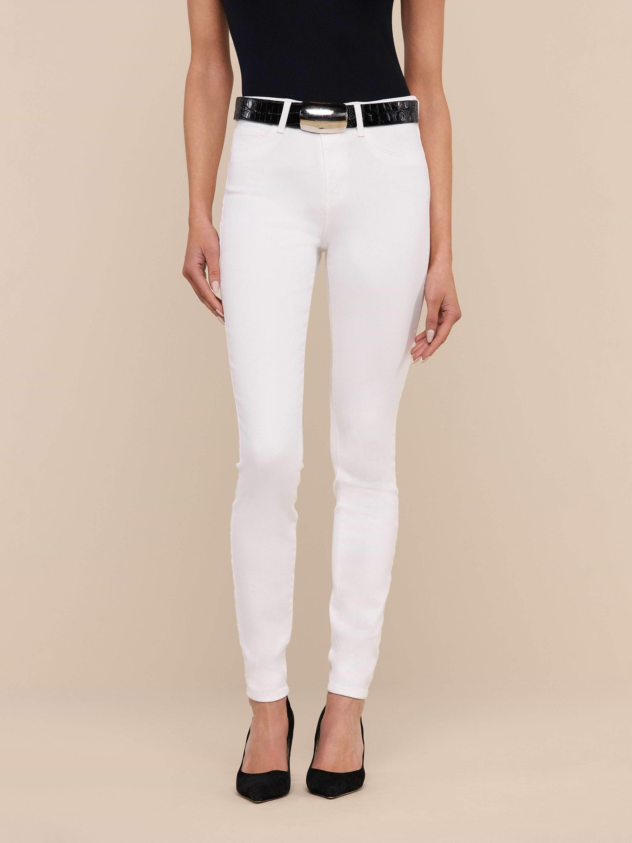 A woman stands against a plain background, wearing a black sleeveless top, the Marguerite Skinny Jean in white with a black belt, and black pointed-toe shoes. Only her body from the shoulders down is visible.