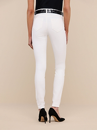 Person standing against a plain background, shown from the waist down, wearing the Marguerite Skinny Jean in white high-recovery fabric, paired with a black top, a white belt, and black high-heeled shoes.