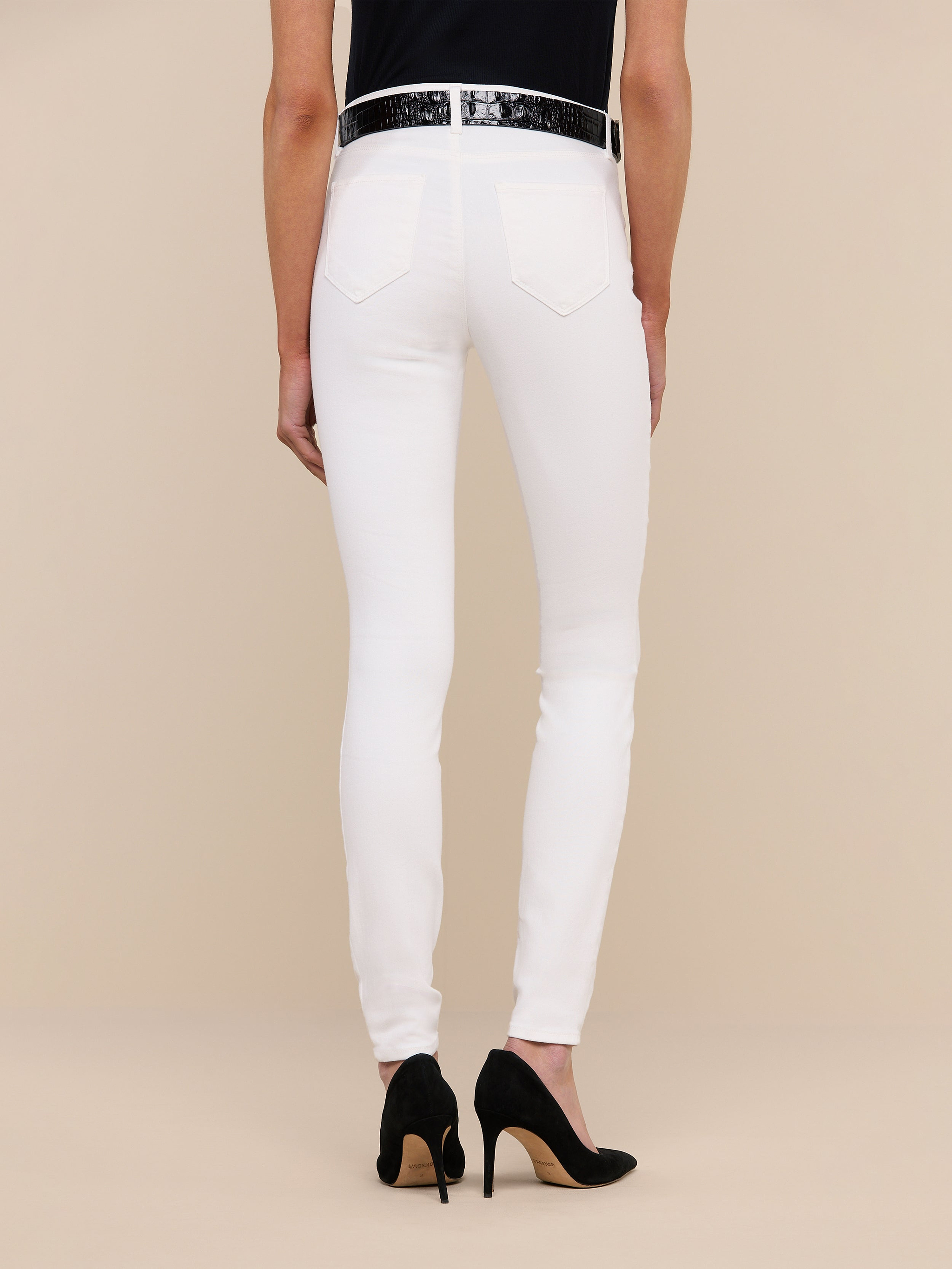 Person standing against a plain background, shown from the waist down, wearing the Marguerite Skinny Jean in white high-recovery fabric, paired with a black top, a white belt, and black high-heeled shoes.