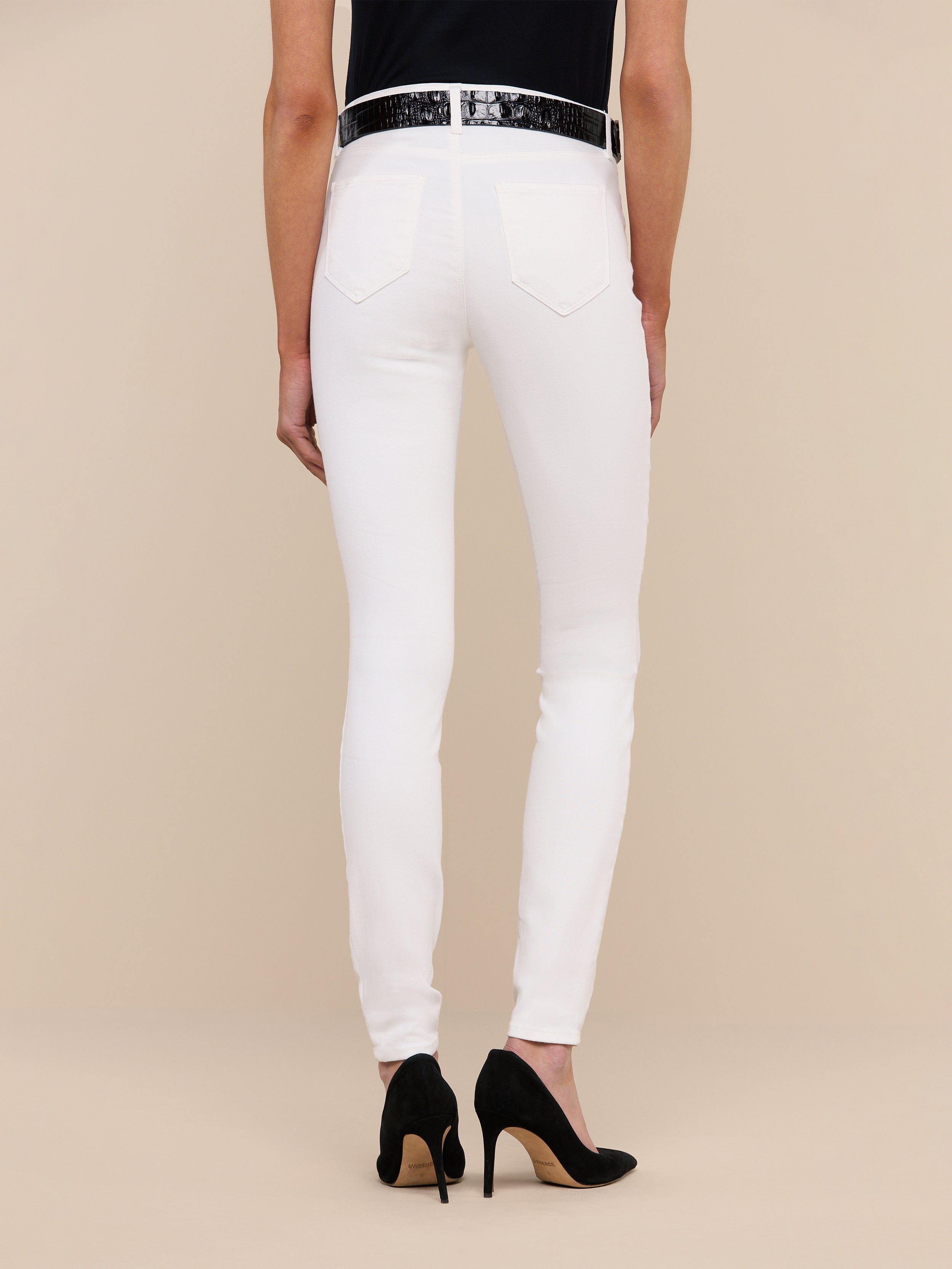 Person standing against a plain background, shown from the waist down, wearing the Marguerite Skinny Jean in white high-recovery fabric, paired with a black top, a white belt, and black high-heeled shoes.
