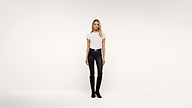 Woman standing against a plain white background, wearing a white short-sleeved shirt, black leather pants, and black boots. She has long blonde hair and a neutral expression.