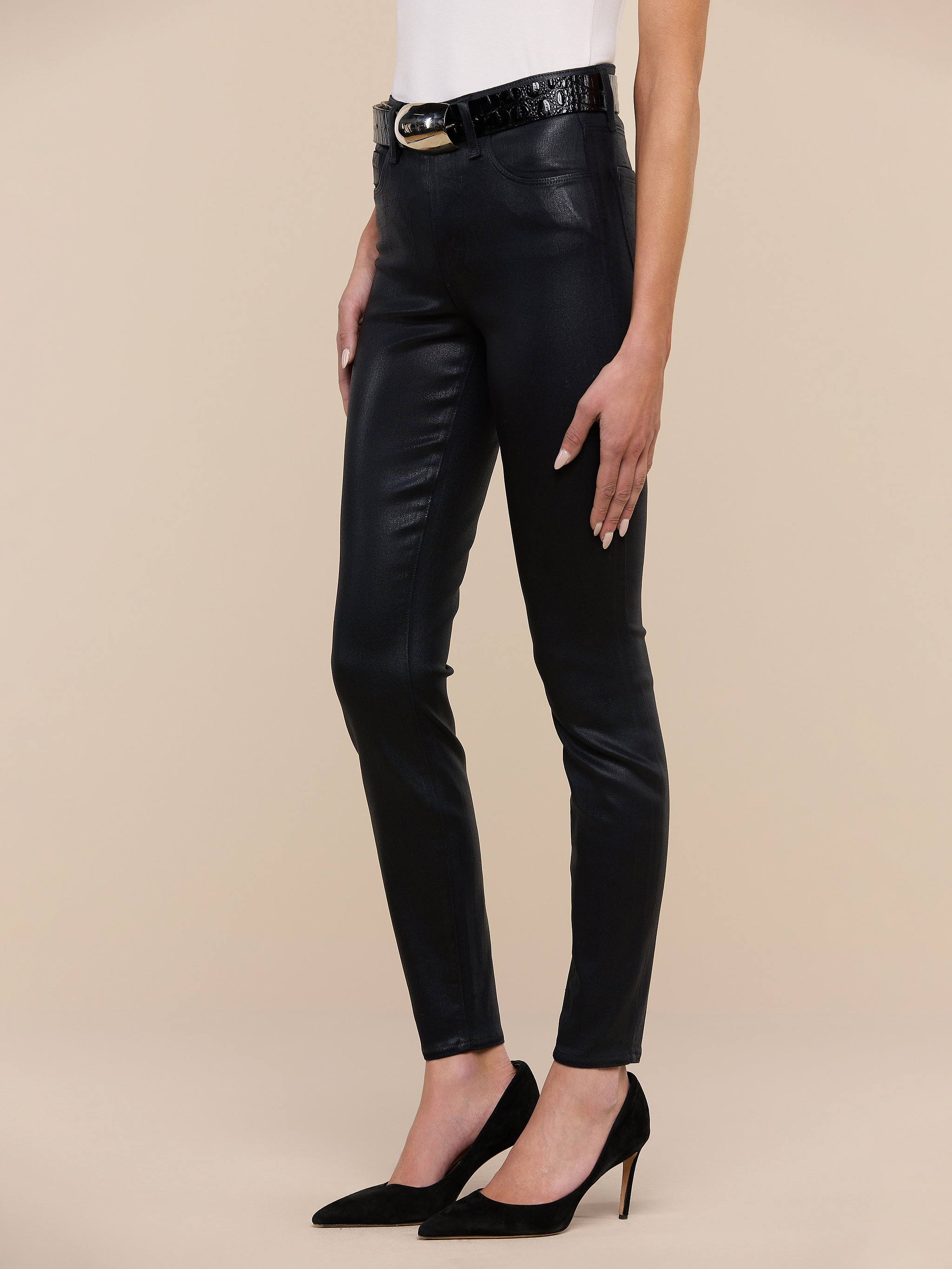 Wearing the Marguerite Coated Skinny Jean in black, a person styled a white top, a black belt, and black high-heeled shoes while standing against a plain white background.