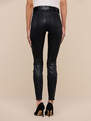 A person stands with their back to the camera, wearing a white top, Marguerite Coated Skinny Jean in black, a black belt, and black high-heeled shoes against a beige background.