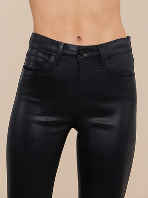 A close-up of a person wearing high-waisted, shiny black Marguerite Coated Skinny Jeans in stretch denim, highlighting the waistband, button, and upper thighs against a beige background with a visible midriff.