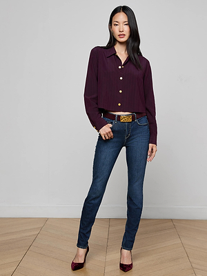 A woman poses on a wood floor in front of a light gray wall, wearing L'AGENCE Marguerite Skinny Jean in stretch denim, a dark purple button-up shirt, wide belt, and burgundy heels.