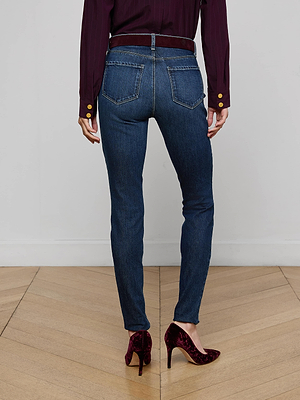 A person stands on a wooden floor facing away, wearing a long-sleeve burgundy shirt, L'AGENCE Marguerite Skinny Jean in stretch denim with back pockets, a belt, and burgundy velvet high-heeled shoes.
