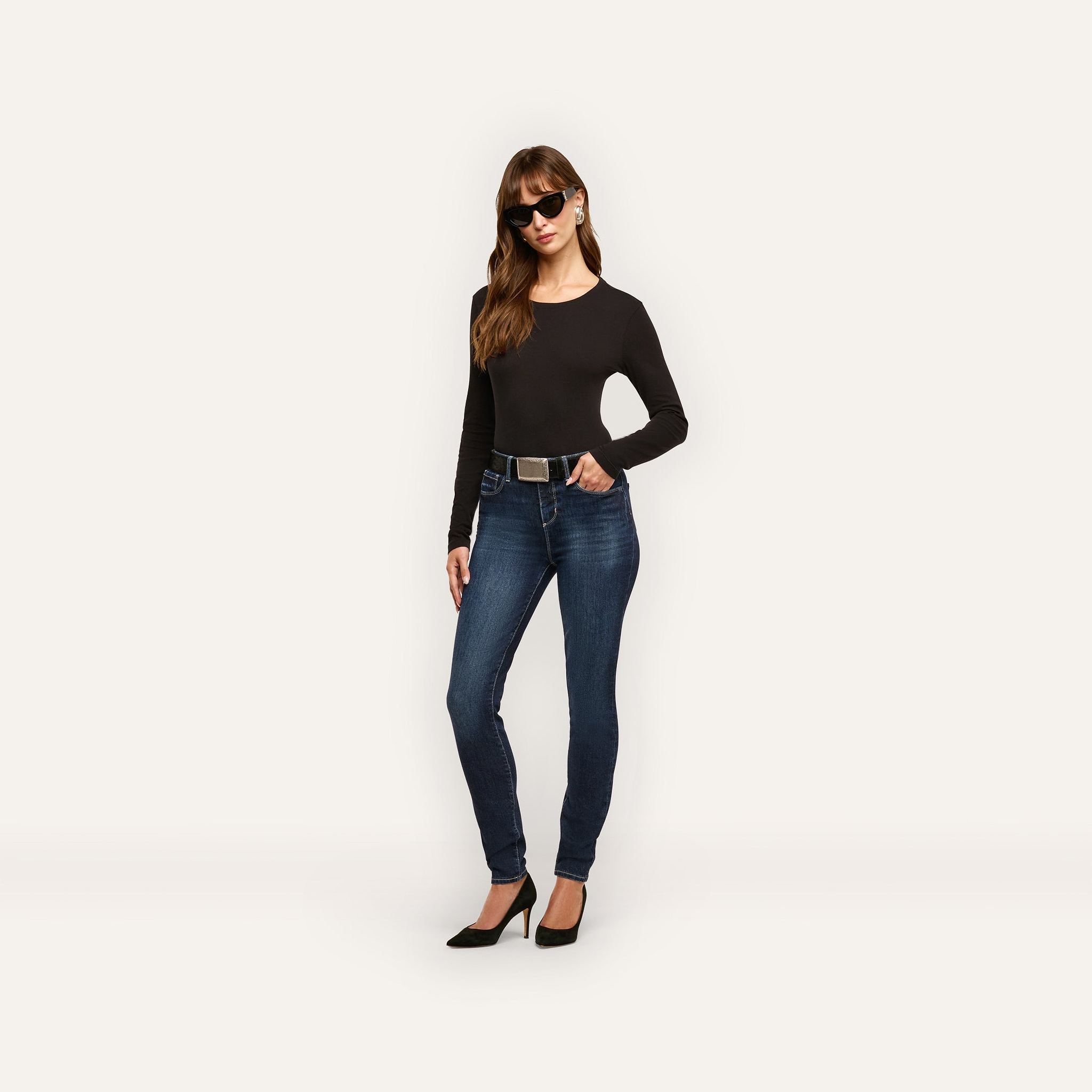 A woman wears a long-sleeved black shirt, Marguerite Skinny Jean high-rise skinny jeans with a belt, and black heels.