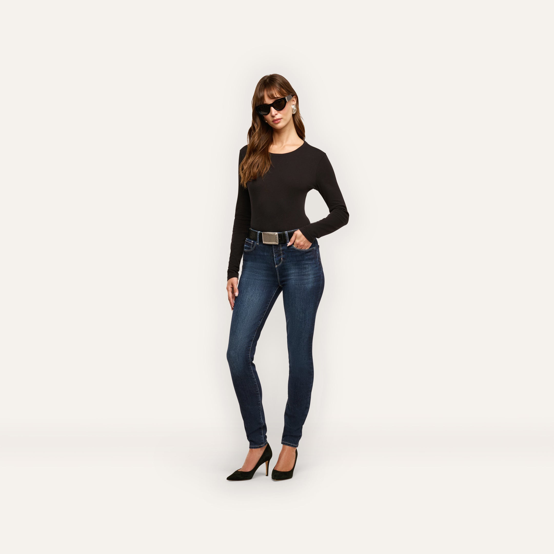 A woman wears a long-sleeved black shirt, Marguerite Skinny Jean high-rise skinny jeans with a belt, and black heels.