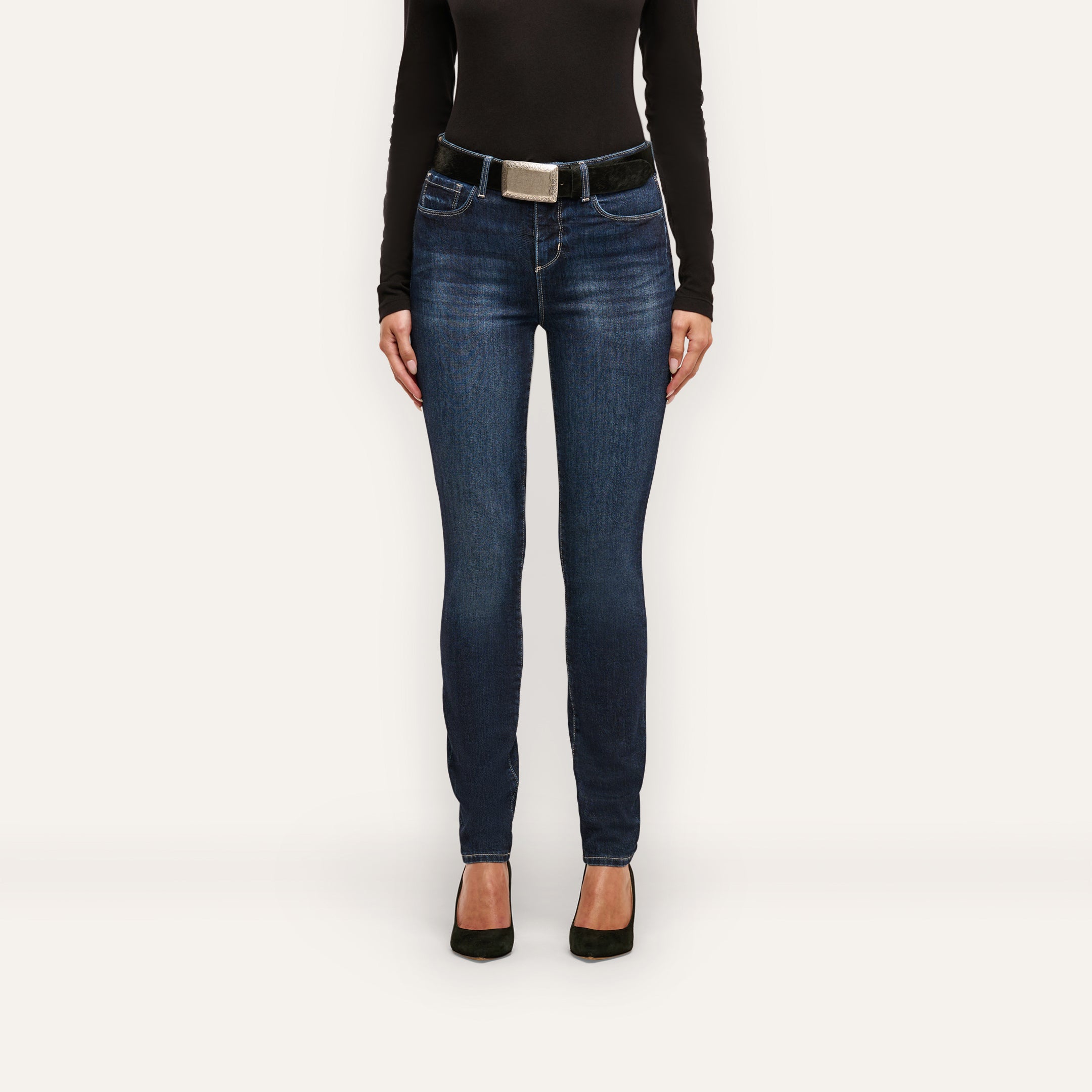 Marguerite Skinny Jean in dark blue with a high-rise fit, styled with a black belt featuring a silver buckle, a black long-sleeve top, and black heels.