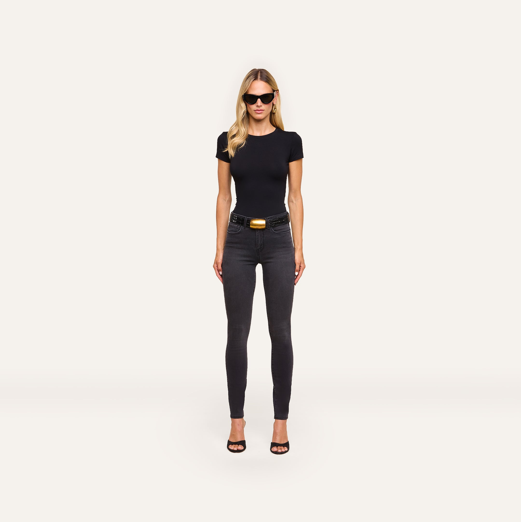 A woman wearing sunglasses, a fitted black t-shirt, Marguerite Skinny Jean with a 30-inch inseam, a black belt with a gold buckle, and black heels stands against a plain light background.