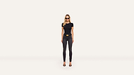A woman in sunglasses, a black t-shirt, dark skinny jeans, and black heels with a gold-buckled belt stands confidently against a minimal, light background.