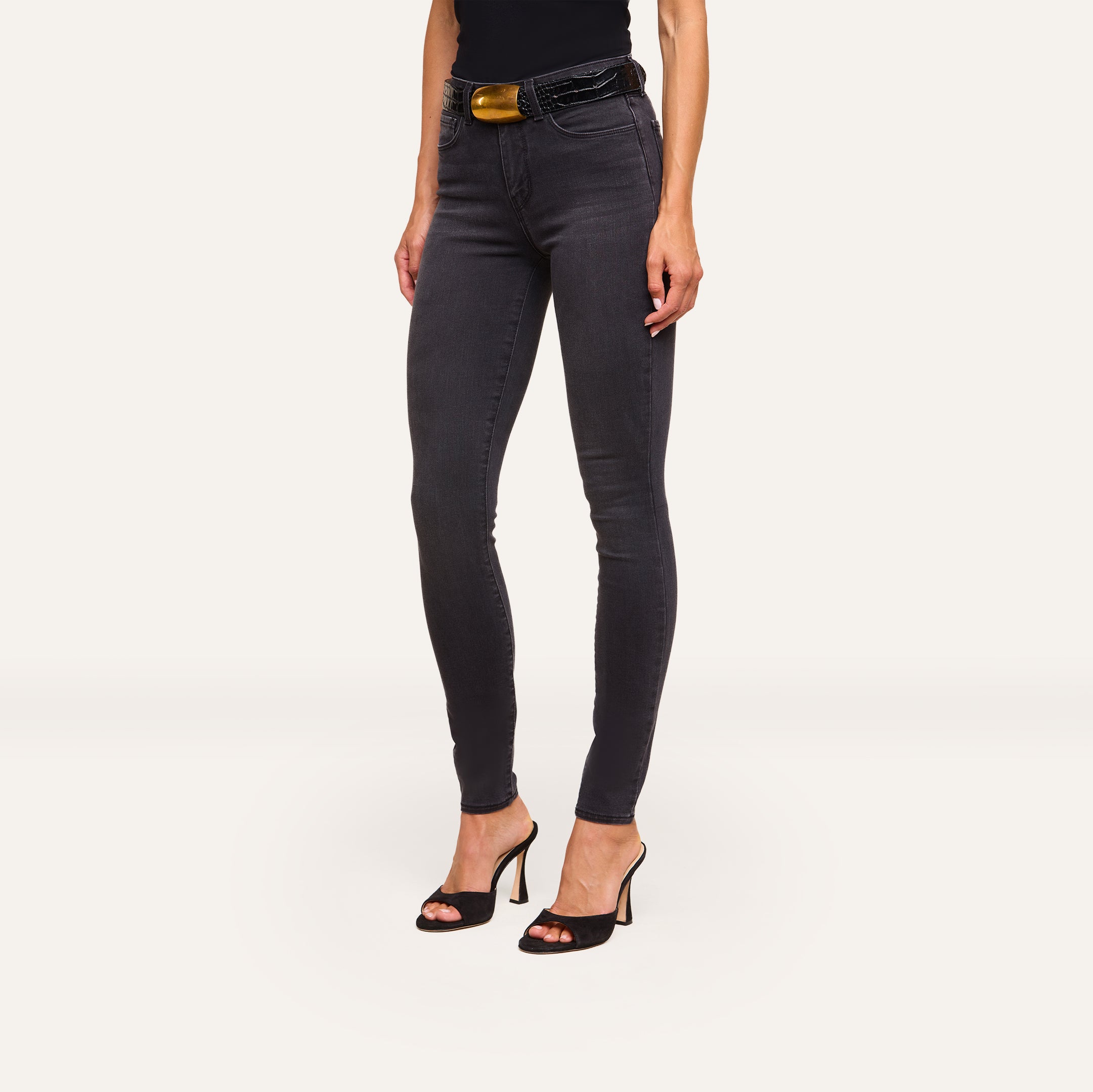 A woman wearing Marguerite Skinny Jean in black, a matching top, a black belt with a gold buckle, and black high-heeled sandals stands against a plain white background.