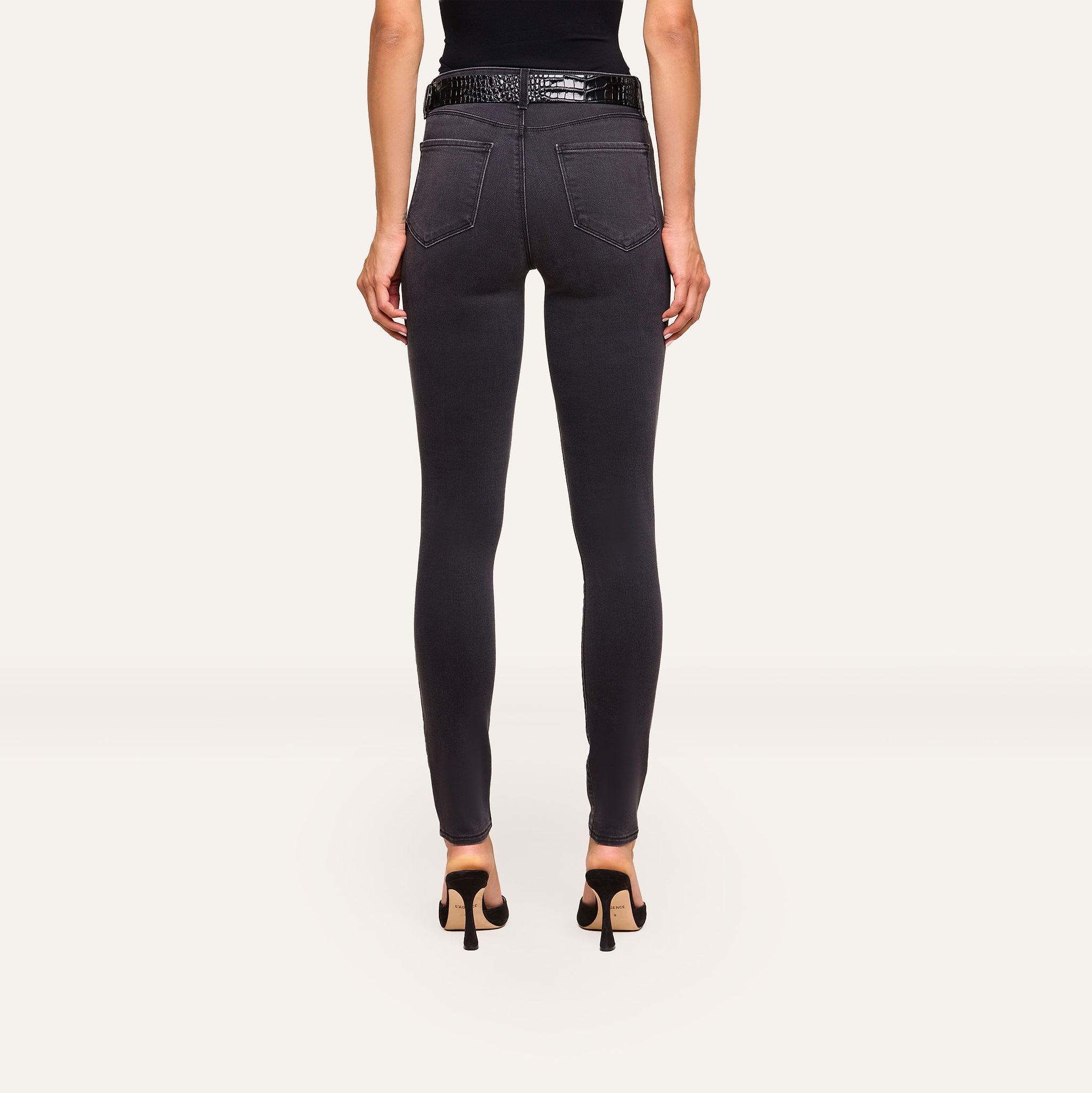 A woman faces away, wearing the Marguerite Skinny Jean in black with a 30-inch inseam and a black sleeveless top, paired with high heels, against a plain light background.