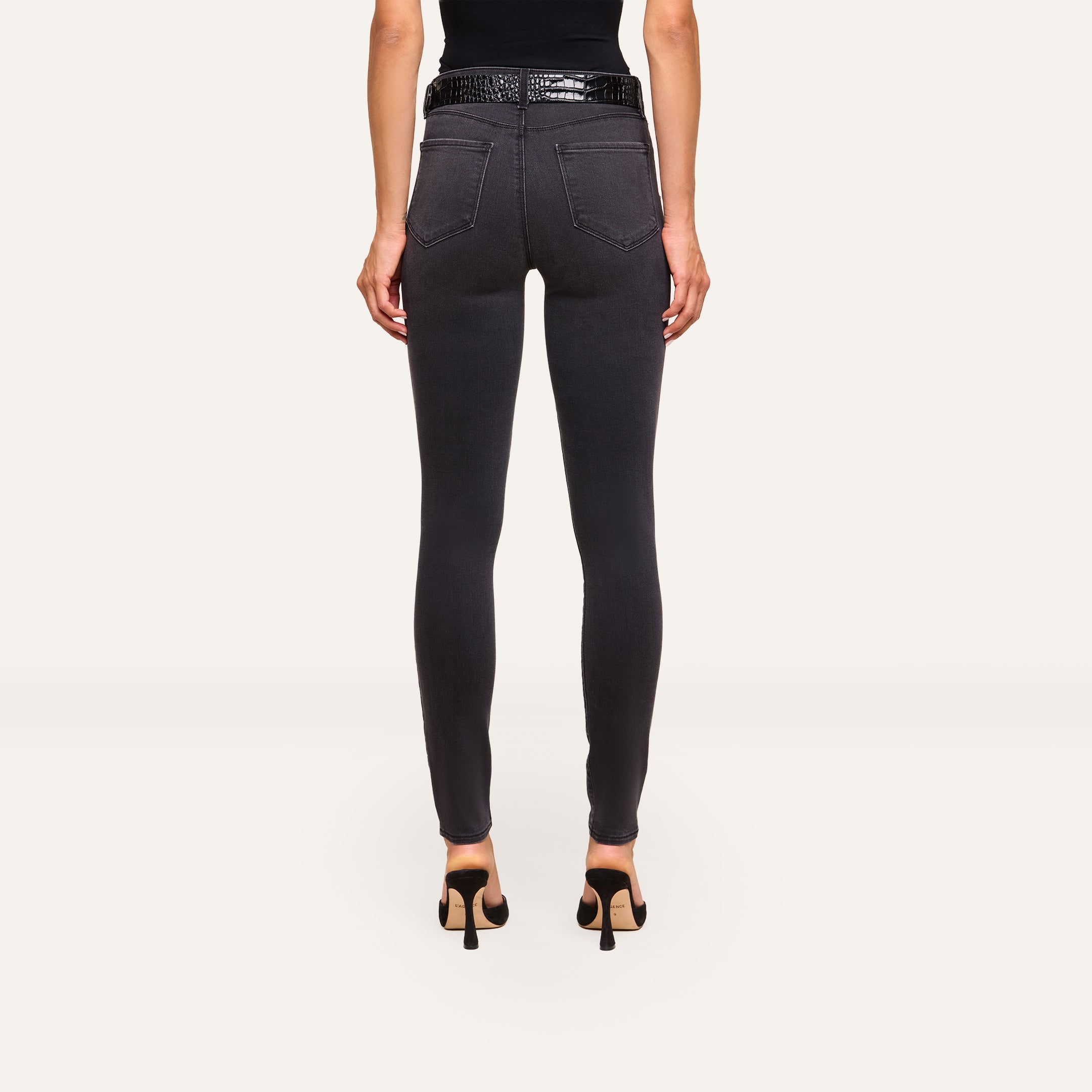 A woman faces away, wearing the Marguerite Skinny Jean in black with a 30-inch inseam and a black sleeveless top, paired with high heels, against a plain light background.