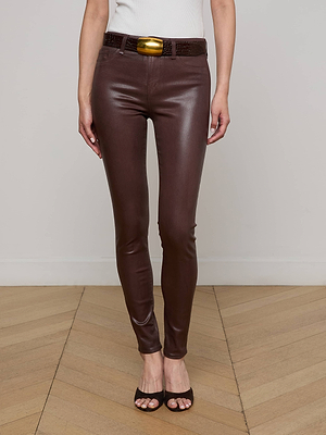 A person stands on a wooden floor wearing L'AGENCE Marguerite Coated Skinny Jeans in chocolate brown, paired with a wide dark belt with a gold buckle, black high-heeled sandals, and a white sleeveless top; only the lower half is shown.