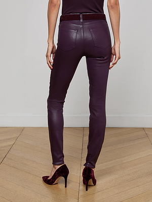 A person stands with their back to the camera, wearing L'AGENCE Marguerite Coated Skinny Jeans in dark purple and matching high-heeled shoes on a light wooden floor.