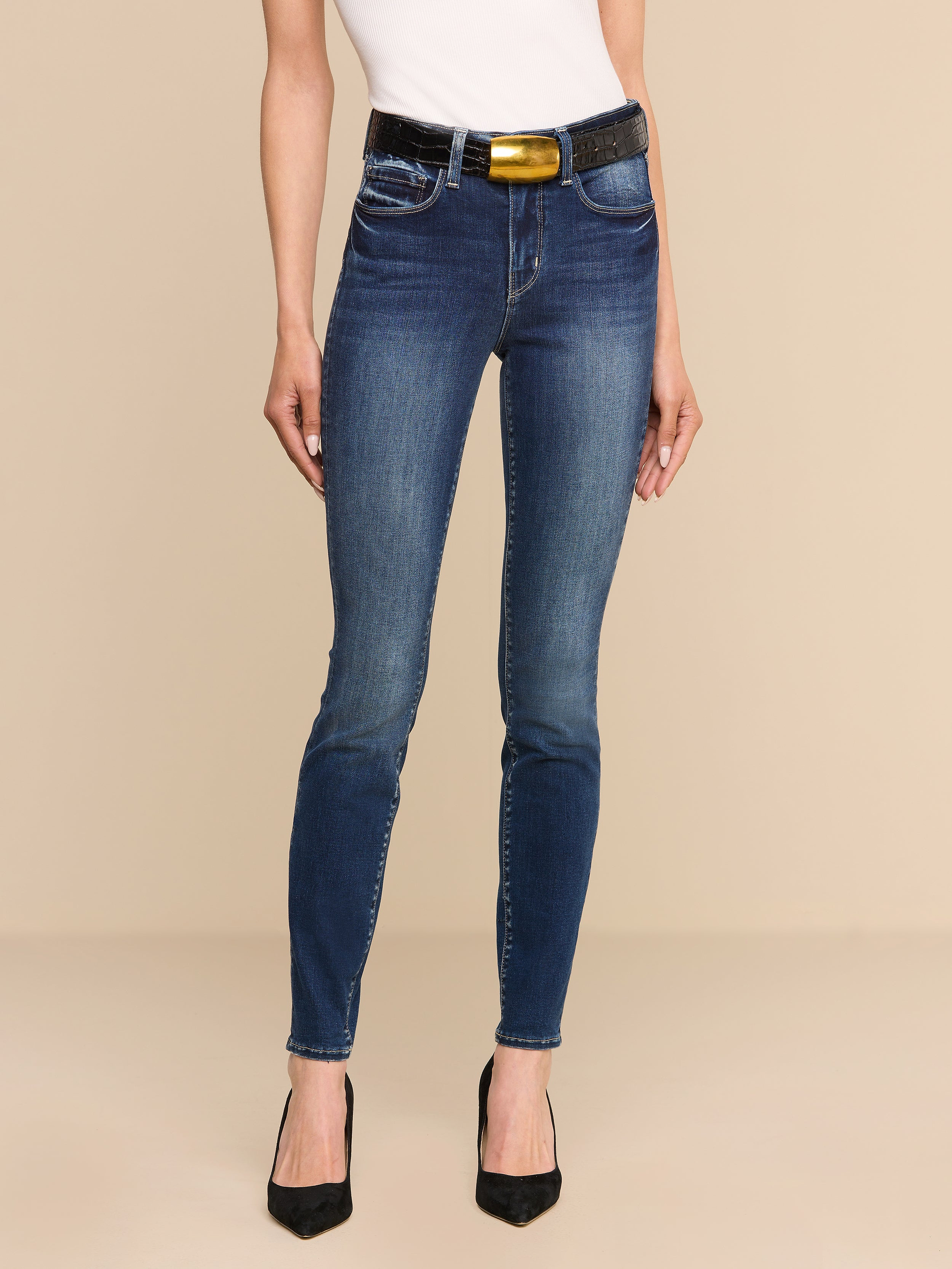 A person wearing a white top, the Marguerite Skinny Jean with a black belt, and black high heels stands against a plain white background. The Marguerite Skinny Jean is crafted from premium stretch denim for added comfort.