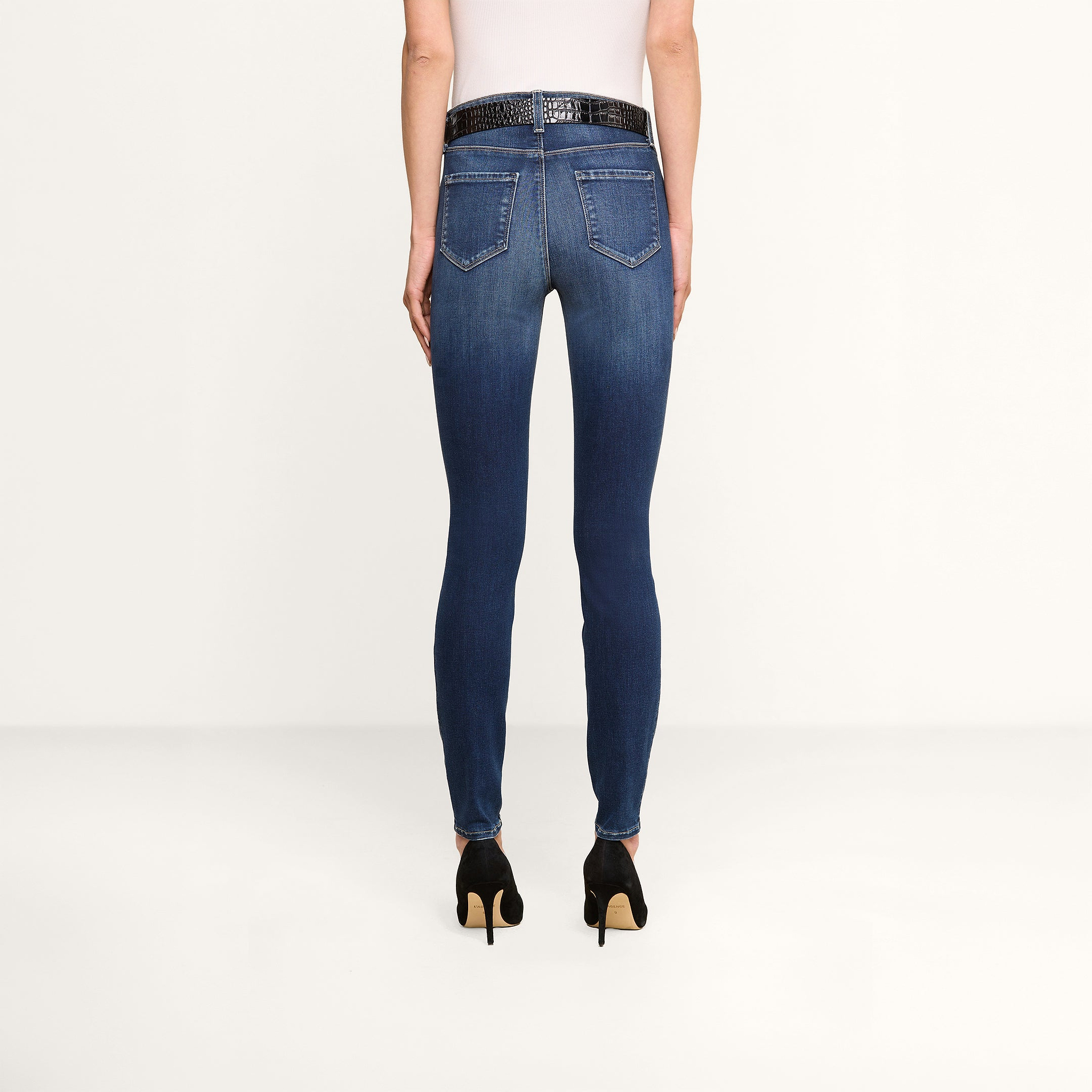 A person wearing a white top, the Marguerite Skinny Jean made from premium stretch denim, a black belt, and black high-heeled shoes is standing facing away from the camera against a plain white background.