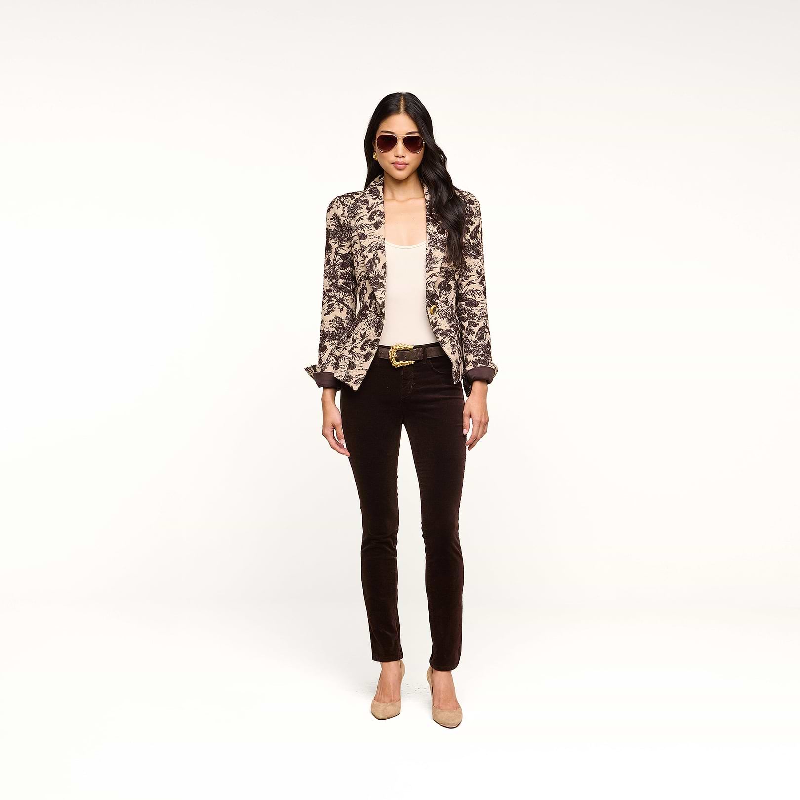 A woman stands against a white background, facing forward with her hands by her sides. She wears sunglasses, a patterned blazer over a light top, the Marguerite Velvet Skinny Jean with a belt, and beige heels.