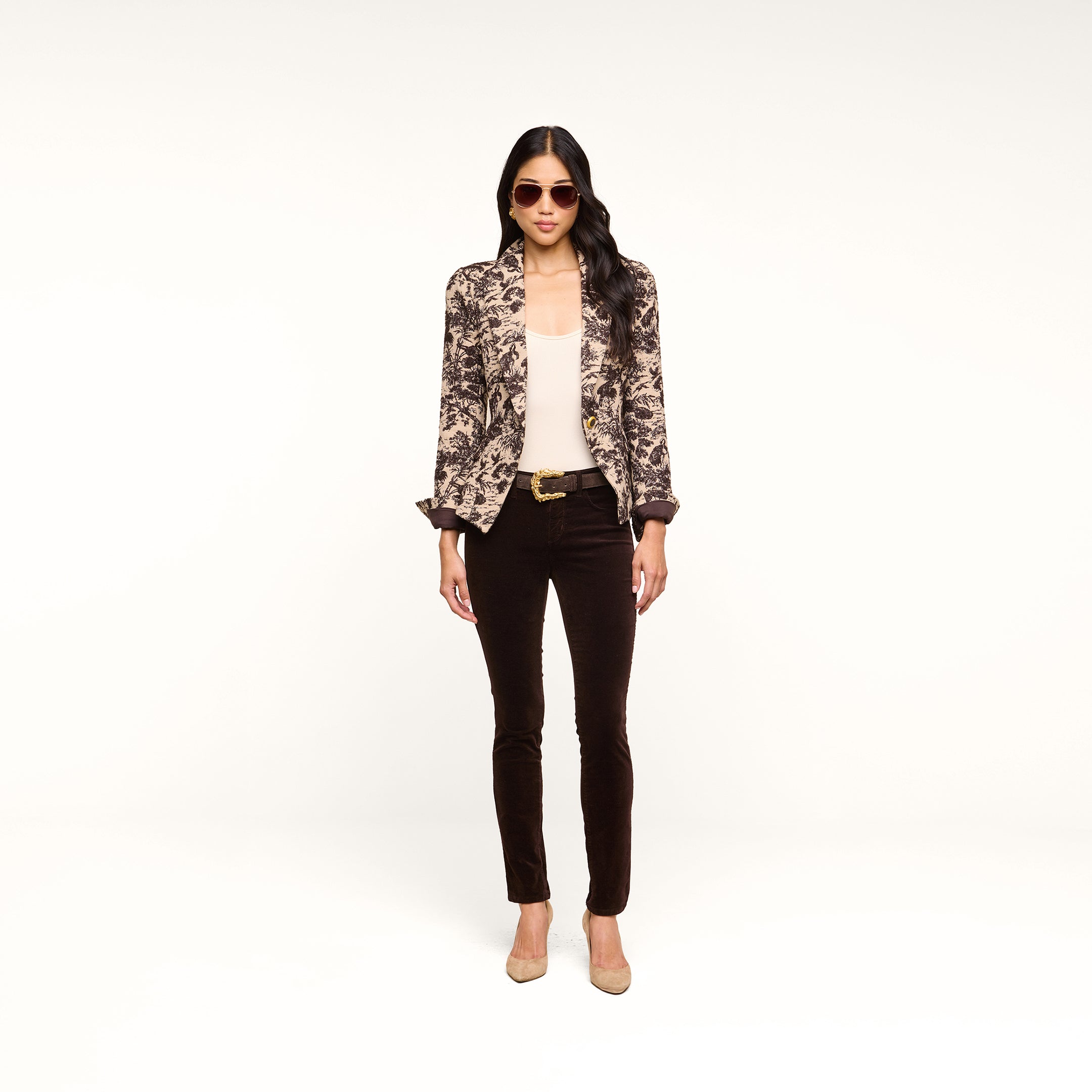 A woman stands against a white background, facing forward with her hands by her sides. She wears sunglasses, a patterned blazer over a light top, the Marguerite Velvet Skinny Jean with a belt, and beige heels.