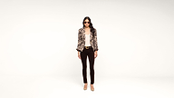 A woman stands against a white background wearing sunglasses, a patterned blazer, a light top, dark pants, and beige heels.