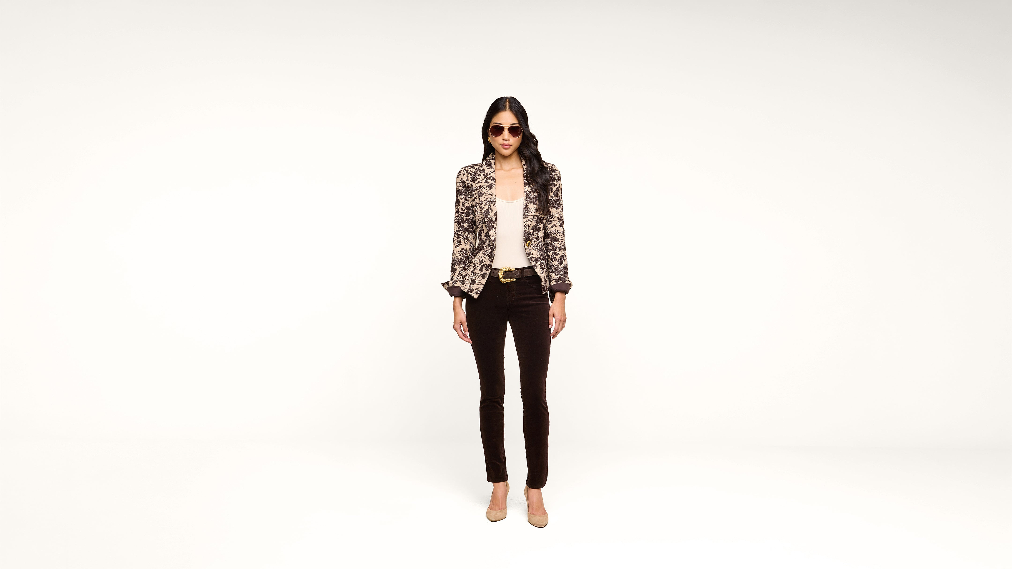 A woman stands against a white background wearing sunglasses, a patterned blazer, a light top, dark pants, and beige heels.