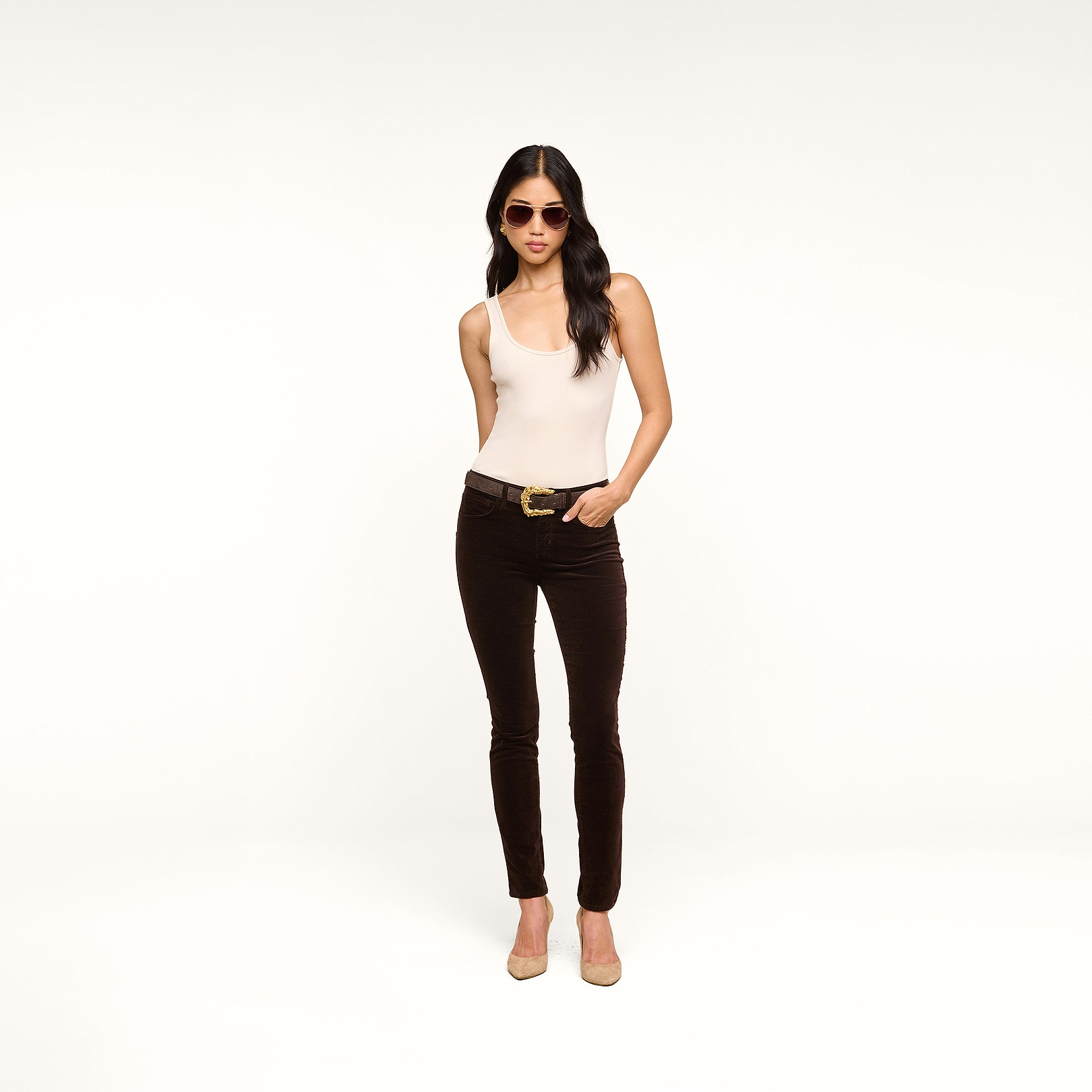 A woman stands with one hand in her pocket against a plain white background, wearing sunglasses, a beige sleeveless top, a belt, beige heels, and the Marguerite Velvet Skinny Jean in a dark high-rise stretch style.