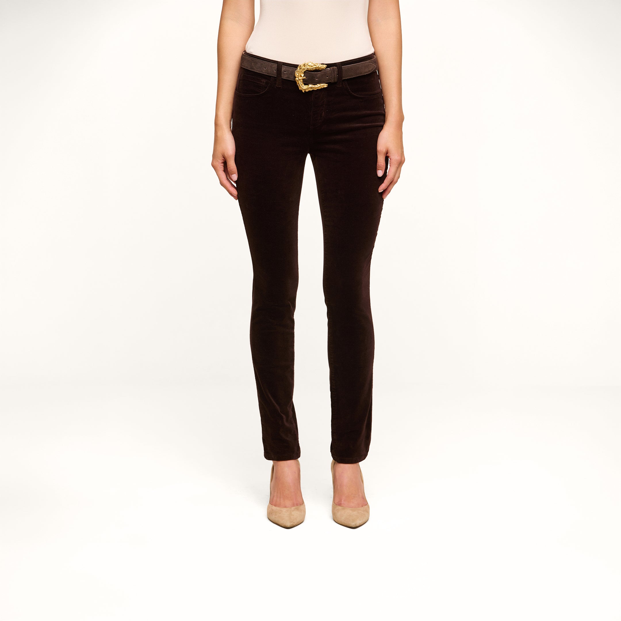 A person stands facing forward, wearing a sleeveless beige top, the Marguerite Velvet Skinny Jean in dark brown with a belt, and beige high-heeled shoes, against a plain white background. Their head is not visible in the image.