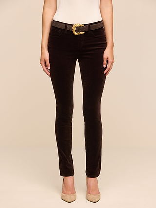 A person wearing the Marguerite Velvet Skinny Jean in dark brown, a white sleeveless top, a black belt with a gold buckle, and beige pointed-toe shoes stands against a plain light background.