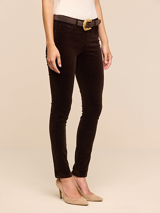 A person wearing the Marguerite Velvet Skinny Jean in dark brown, paired with a white sleeveless top, a black belt with a gold buckle, and beige high heels, standing against a plain beige background.