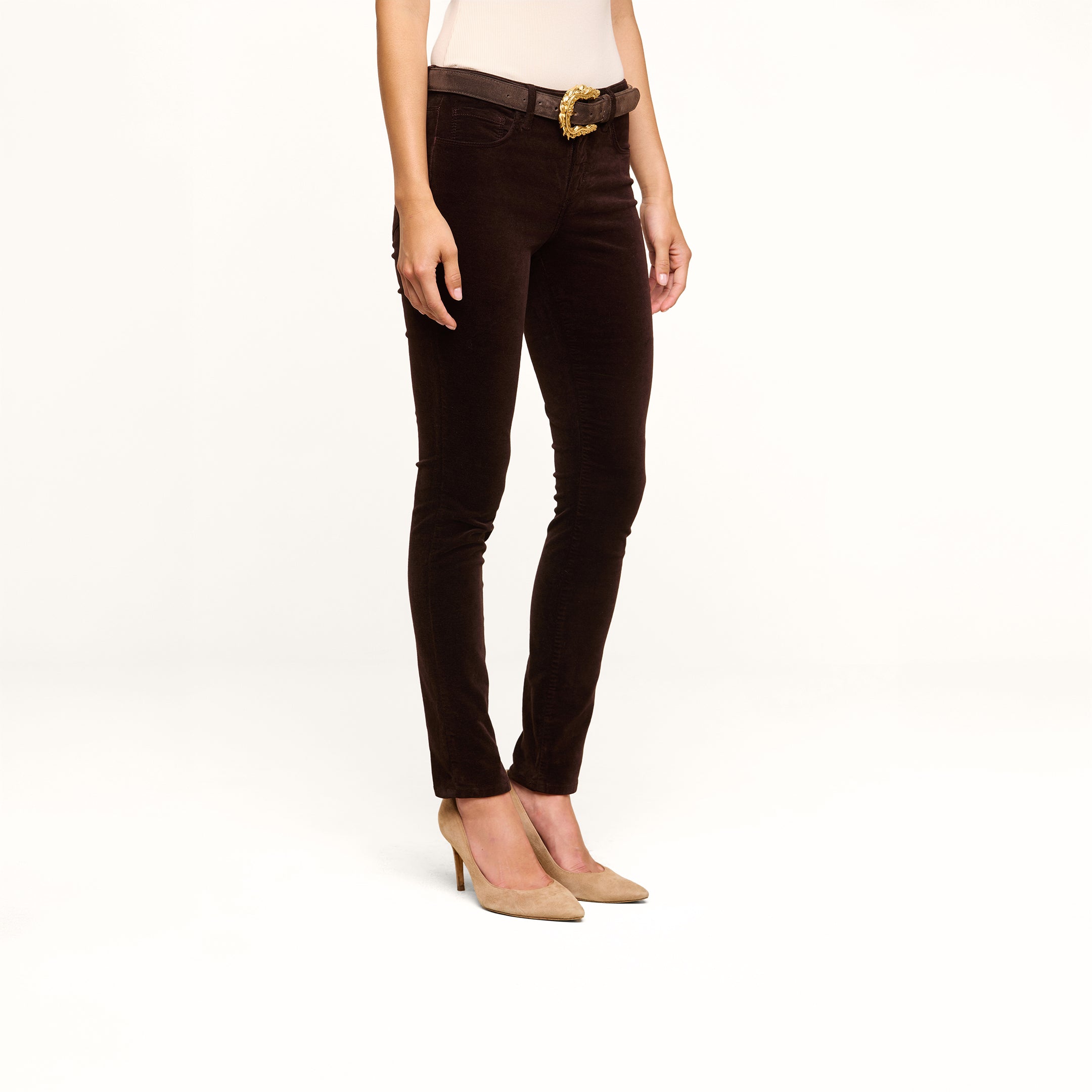 A person wearing the Marguerite Velvet Skinny Jean in dark brown, a beige top, a black belt with a gold buckle, and beige high heels stands against a plain white background.