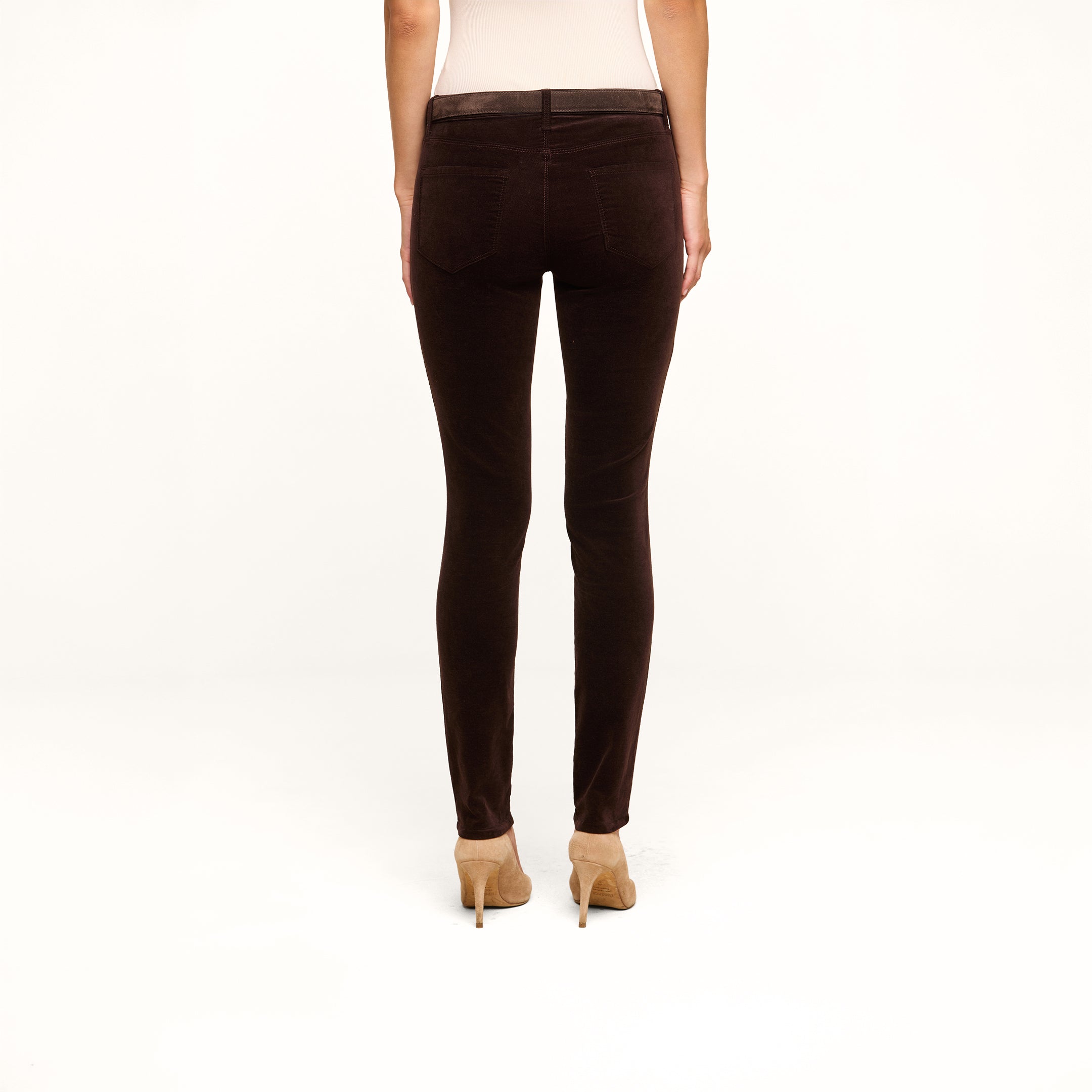A person stands facing away, wearing the Marguerite Velvet Skinny Jean in high rise dark brown stretch velvet and beige high heels, with their arms relaxed by their sides against a plain white background.