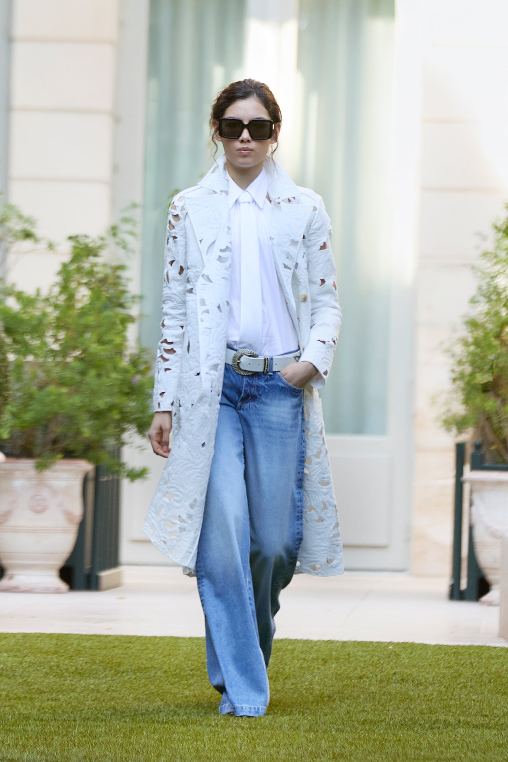 A person in a long, white, textured coat over a white shirt and blue jeans strolls on grass, wearing large sunglasses with hands in pockets. Potted plants and a building with tall windows appear in the background.