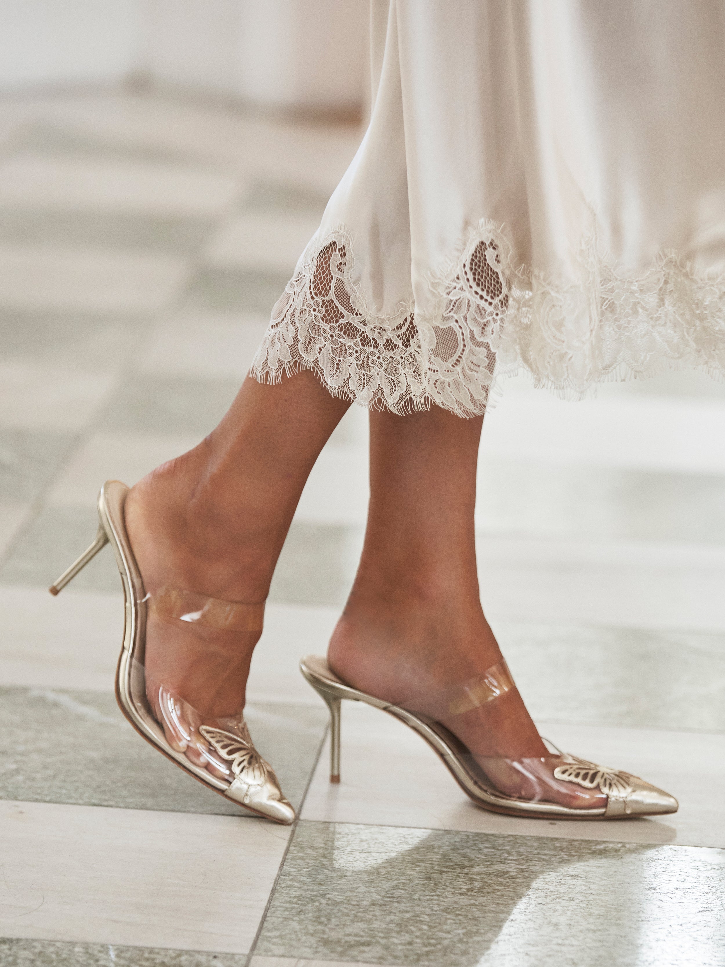 A person wearing a white lace-trimmed dress and clear pointed-toe high heels stands elegantly on a tiled floor, highlighting the delicate lace details.
