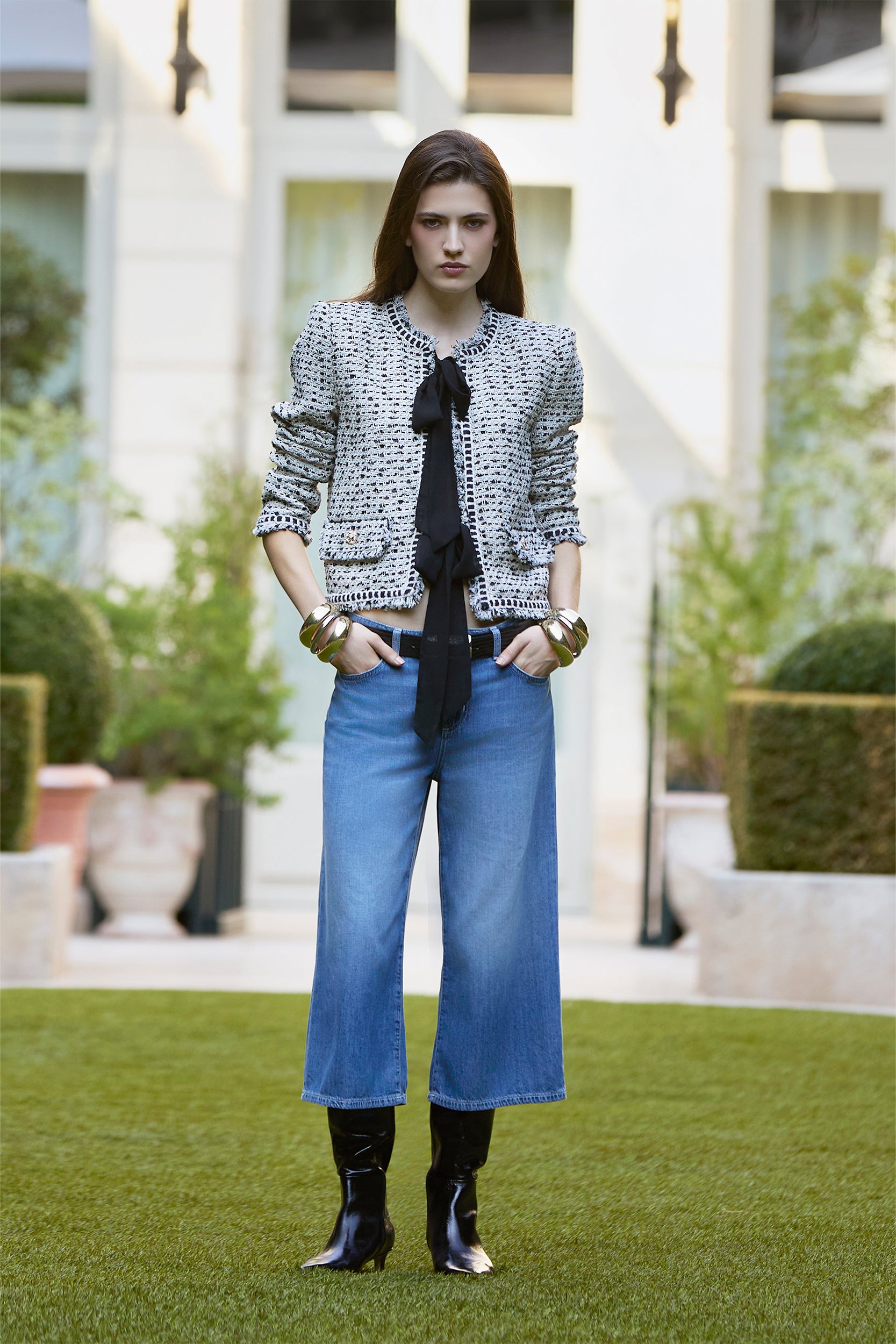 A woman stands on grass wearing a tweed jacket, black blouse with a bow, cropped blue jeans, and black boots. She accessorizes with bracelets against a backdrop of plants and a light-colored building.