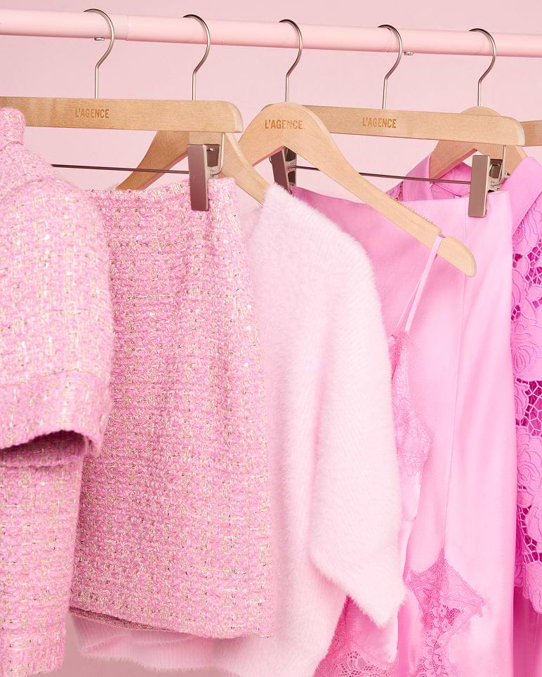 Su un appendiabiti con grucce di legno sono esposti vari capi rosa, tra cui una giacca a maniche corte con trama in rilievo, una gonna abbinata, un top soffice, un abito di seta e un abito di pizzo, tutti in diverse tonalità di rosa.