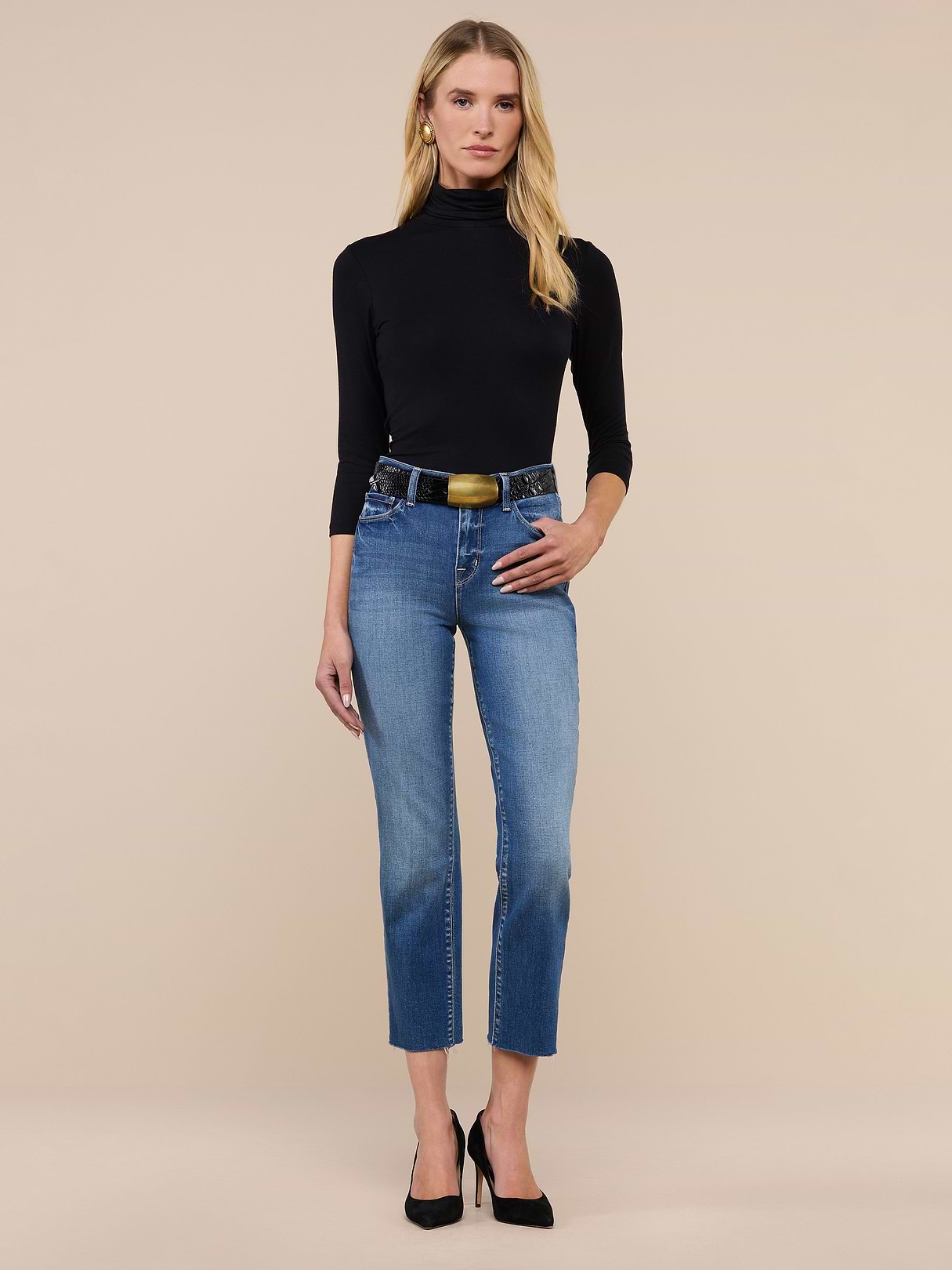 A woman stands against a plain background wearing a black turtleneck, black heels, and the Sada Slim-Leg Cropped Jean—a pair of blue high-rise cropped jeans crafted from premium stretch denim with a black belt. She has long blonde hair and her left hand rests in her pocket.
