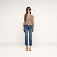 Una donna è in piedi su uno sfondo beige; indossa occhiali da sole, una maglia a maniche lunghe color tortora con tasche sul davanti, jeans Sada Cropped Slim-Leg blu con orlo grezzo, una cintura color cuoio e scarpe con tacco a punta dello stesso colore.