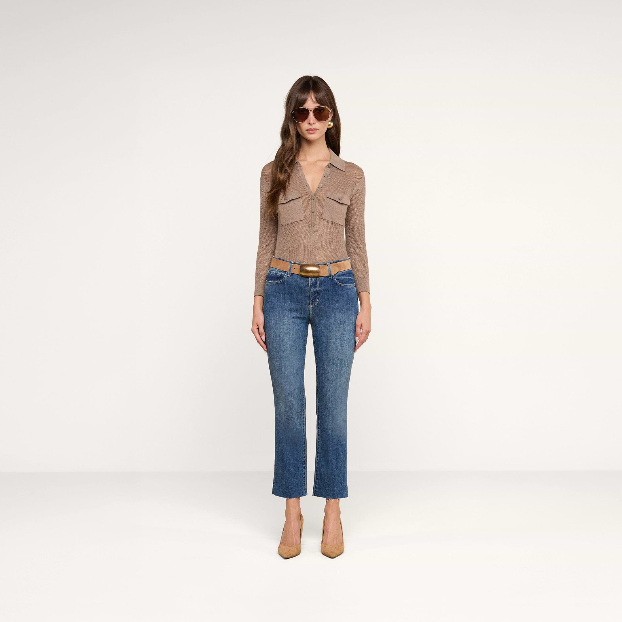 Una donna è in piedi su uno sfondo beige; indossa occhiali da sole, una maglia a maniche lunghe color tortora con tasche sul davanti, jeans Sada Cropped Slim-Leg blu con orlo grezzo, una cintura color cuoio e scarpe con tacco a punta dello stesso colore.