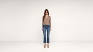 A woman stands in a minimal, white studio wearing sunglasses, a tan long-sleeve top, blue jeans, a brown belt, and beige heels.