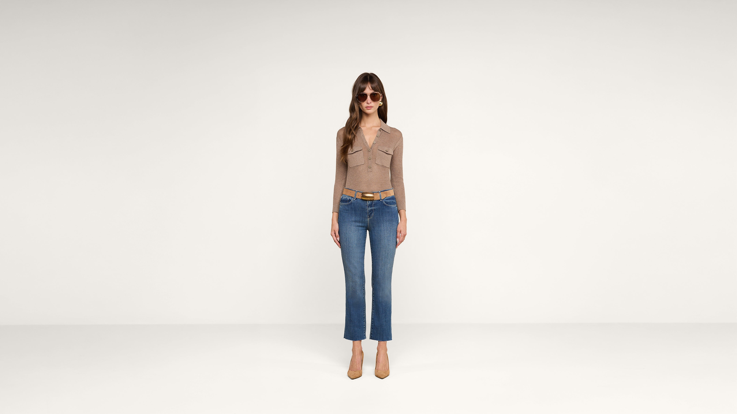 A woman stands in a minimal, white studio wearing sunglasses, a tan long-sleeve top, blue jeans, a brown belt, and beige heels.