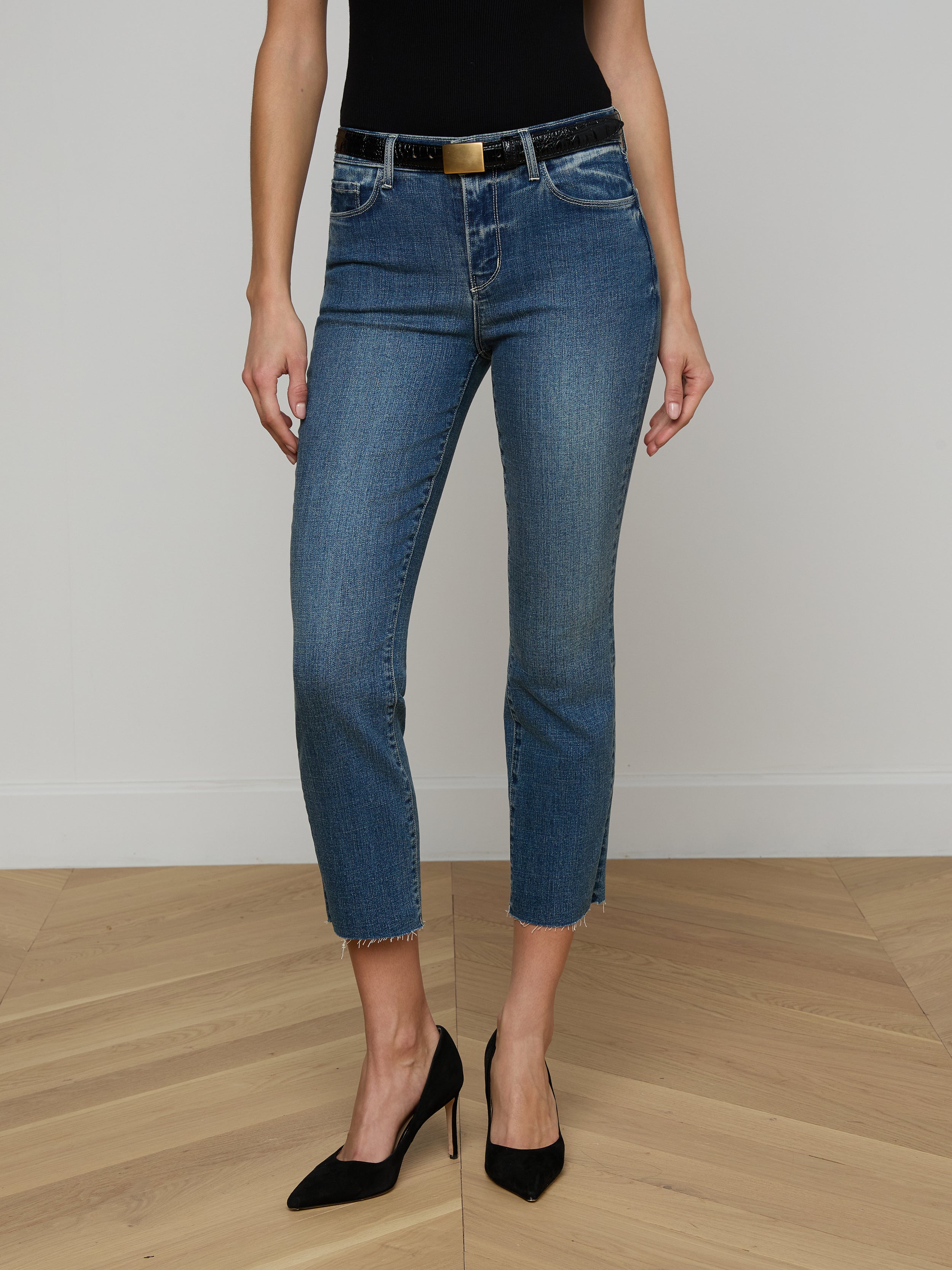 A person wearing a brown long-sleeve top, tan belt, and tan high heels stands against a plain white background, styled in blue Sada Cropped Slim-Leg Jeans with a raw hem.