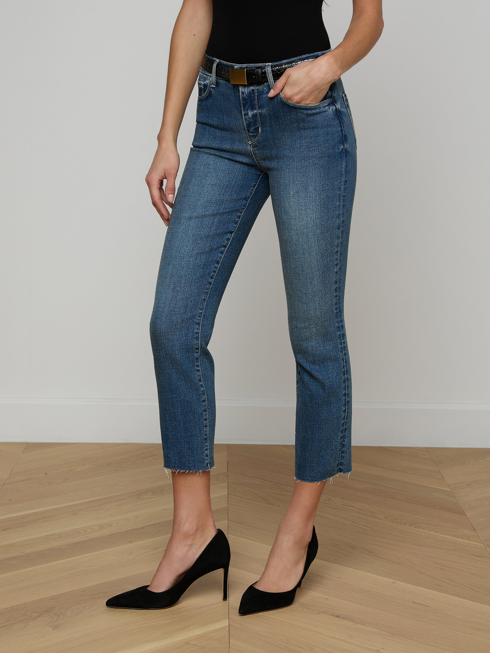 A person wearing a tan long-sleeve top, the Sada Cropped Slim-Leg Jean in blue with a raw hem and a brown belt, and tan high heels stands against a plain white background.