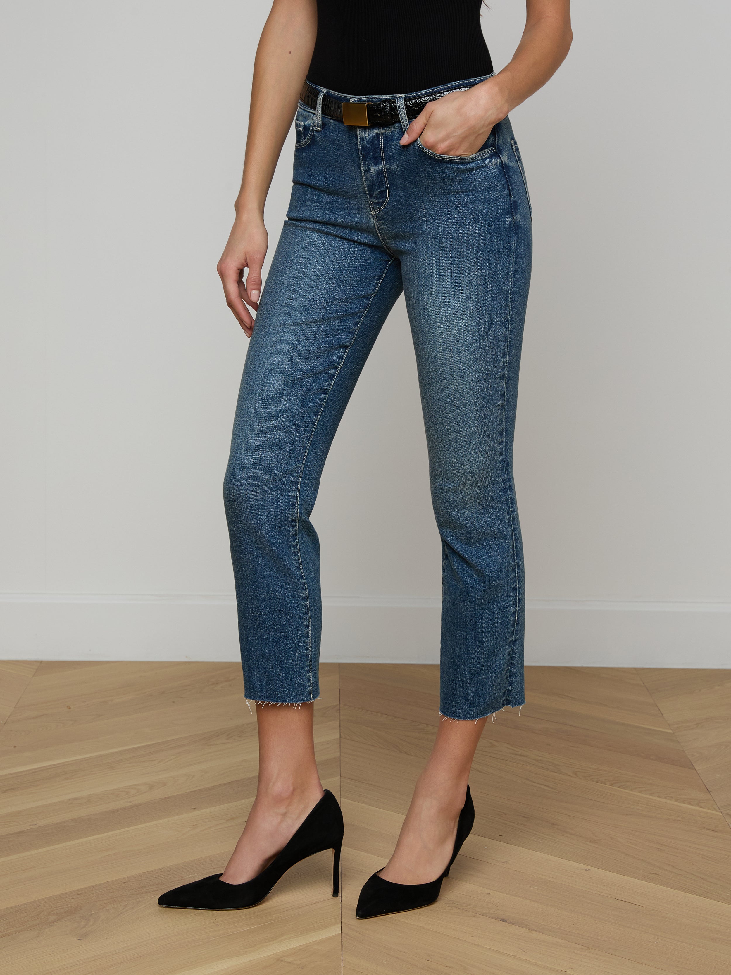 A person wearing a tan long-sleeve top, the Sada Cropped Slim-Leg Jean in blue with a raw hem and a brown belt, and tan high heels stands against a plain white background.