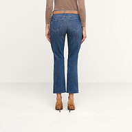A person is shown from the back standing against a plain light background, wearing the Sada Cropped Slim-Leg Jean in denim blue with a raw hem, paired with a brown long-sleeve top and brown heeled shoes.